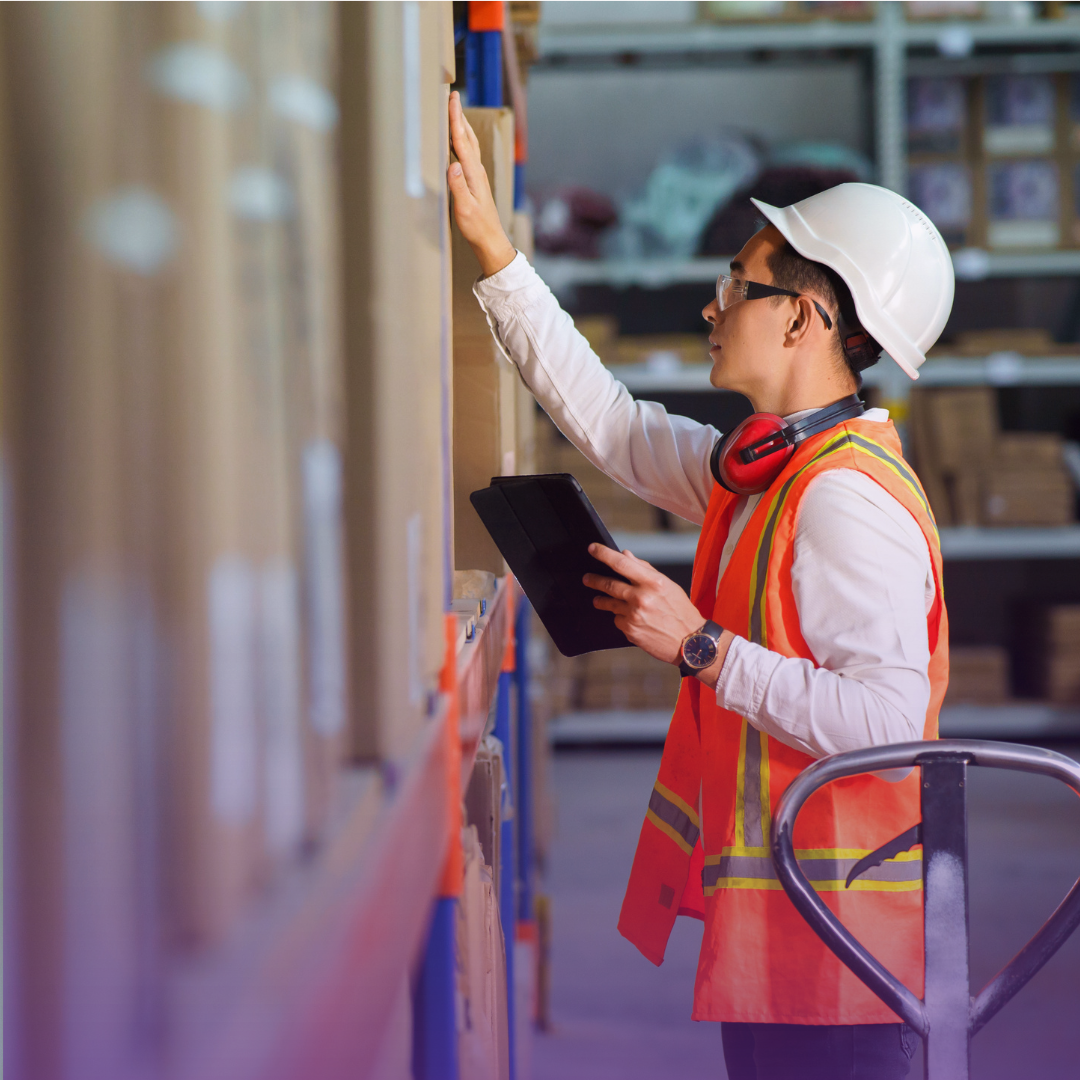 Asian male warehouse worker in safety gear scans boxes on a shelf with a tablet, symbolizing inventory tracking and progress.