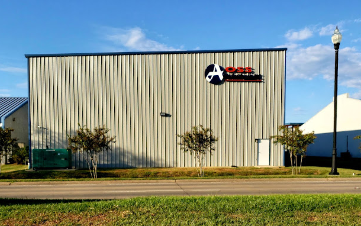 Side view of the AOSS Medical Supply, LLC. industrial building, featuring a prominent blue AOSS logo on its light gray corrugated metal facade, set against a clear blue sky with green grass and trees in the foreground, showcasing a modern facility.