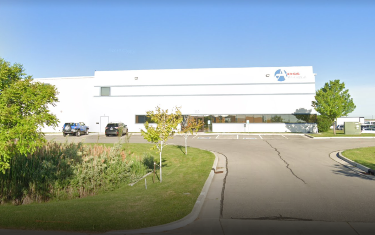 The modern, white exterior of the AOSS Medical Supply, LLC. main facility, featuring a curved driveway, landscaped grounds with trees and shrubs, and parked cars, all under a clear blue sky, highlighting the company's prominent presence.