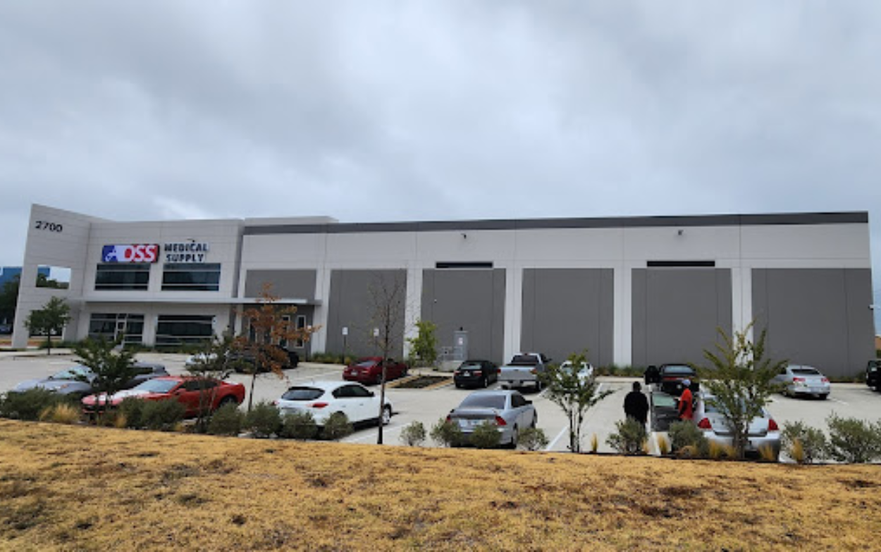 The expansive exterior of a modern AOSS Medical Supply, LLC. distribution center, featuring a two-story white and gray facade with multiple loading bay doors, a large parking lot filled with cars, and landscaped greenery in the foreground under a cloudy sky.