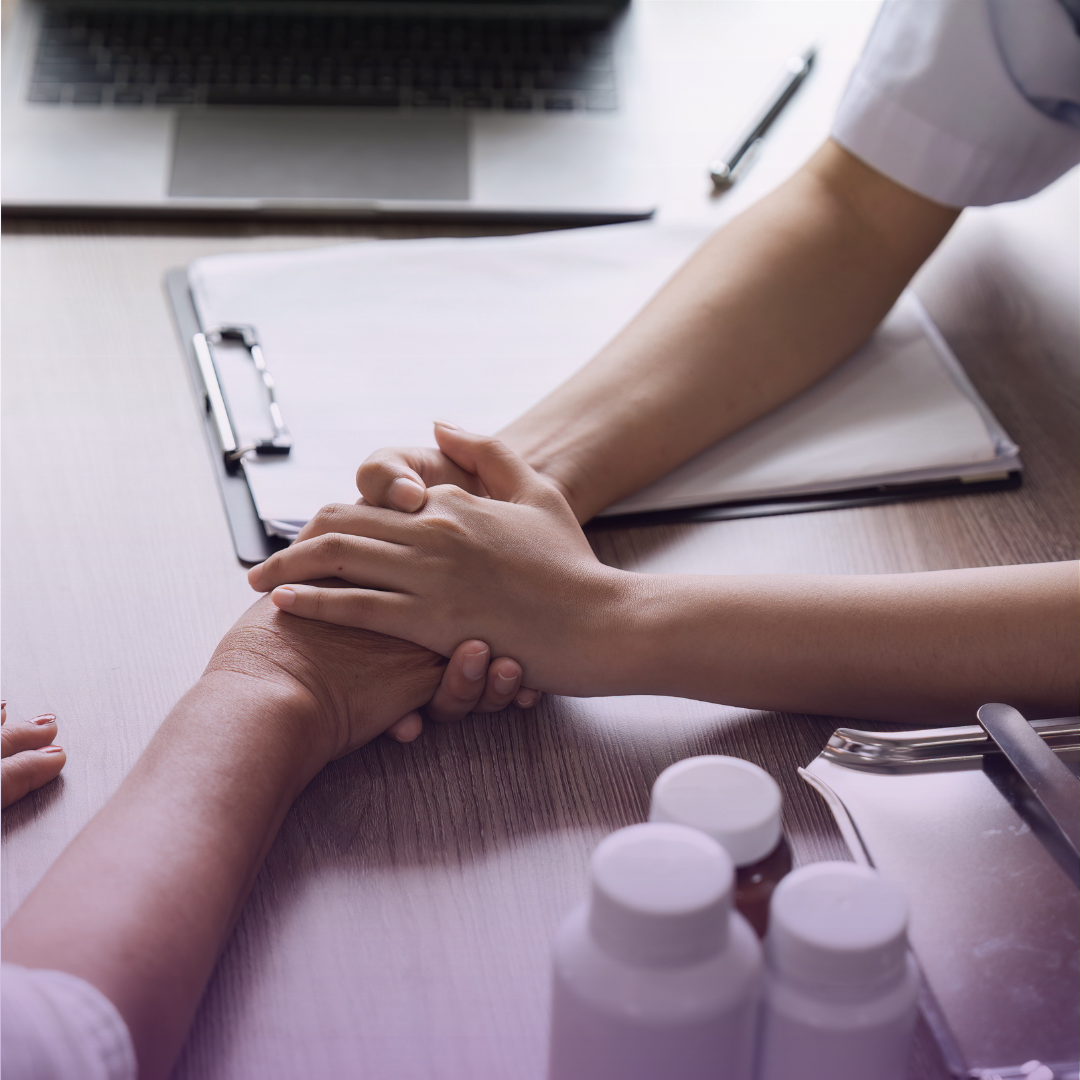 Medical professional gently holds elderly patient's hand on a desk with medical supplies, symbolizing compassionate care.