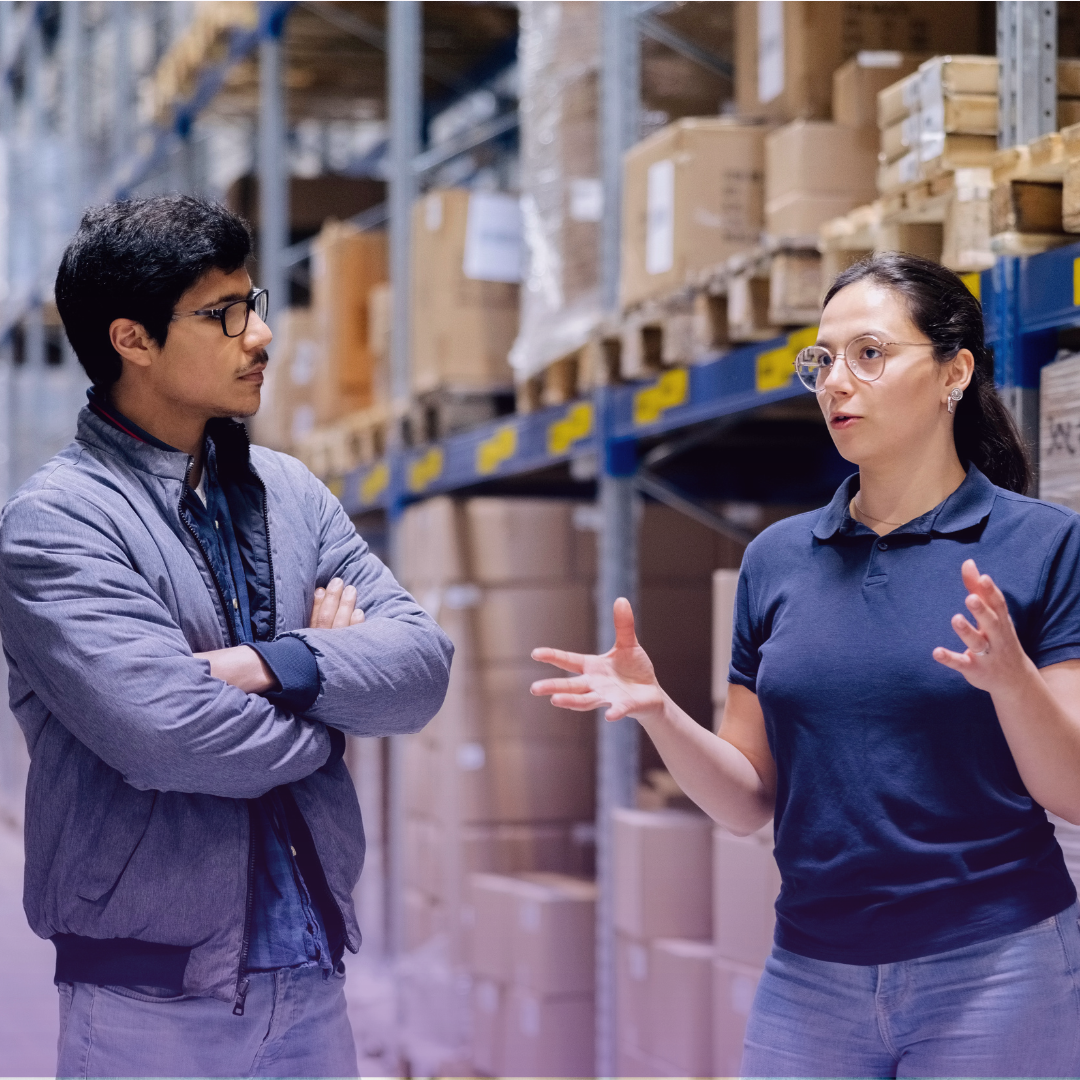 Warehouse supervisor instructs male colleague in an aisle of stacked boxes, symbolizing reliable teamwork.