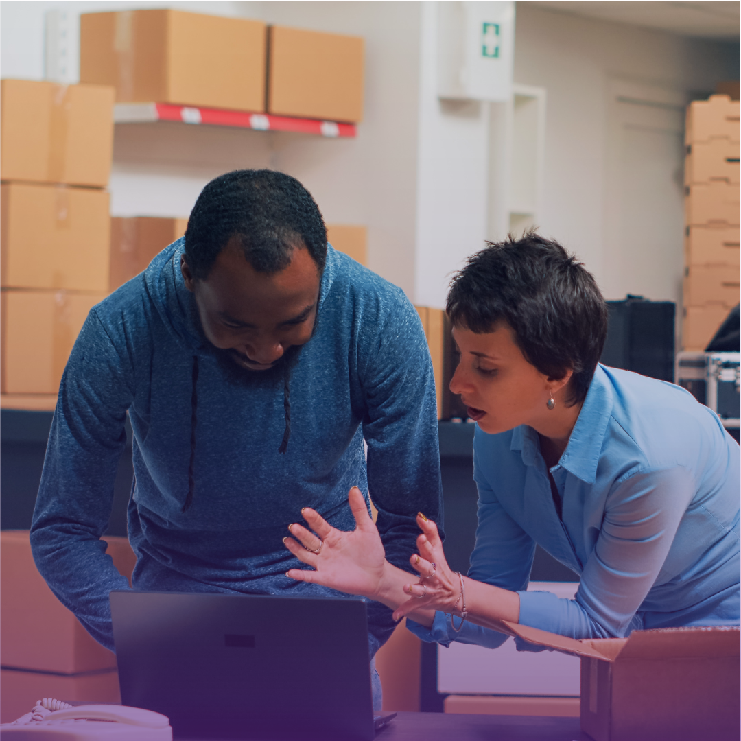 Warehouse professionals collaborate over a laptop with boxes and shelves in the background, symbolizing digital management within the supply chain network.