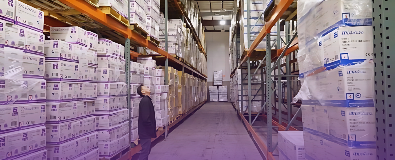 Cover photo for the Distribution page: AOSS Medical Supply CEO Eric Liew stands in a long aisle between towering metal shelves packed densely with white and light blue medical supply boxes, illustrating the vast inventory and organized storage of a modern distribution center.