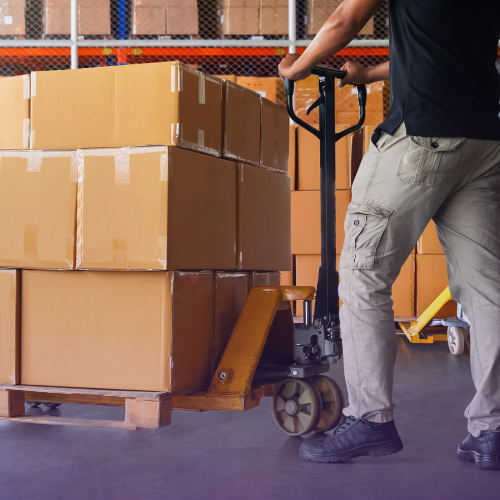 A warehouse worker in cargo pants and a dark shirt pushes a yellow pallet jack loaded with stacks of brown cardboard boxes across a concrete floor, with rows of shelves filled with inventory visible in the background.