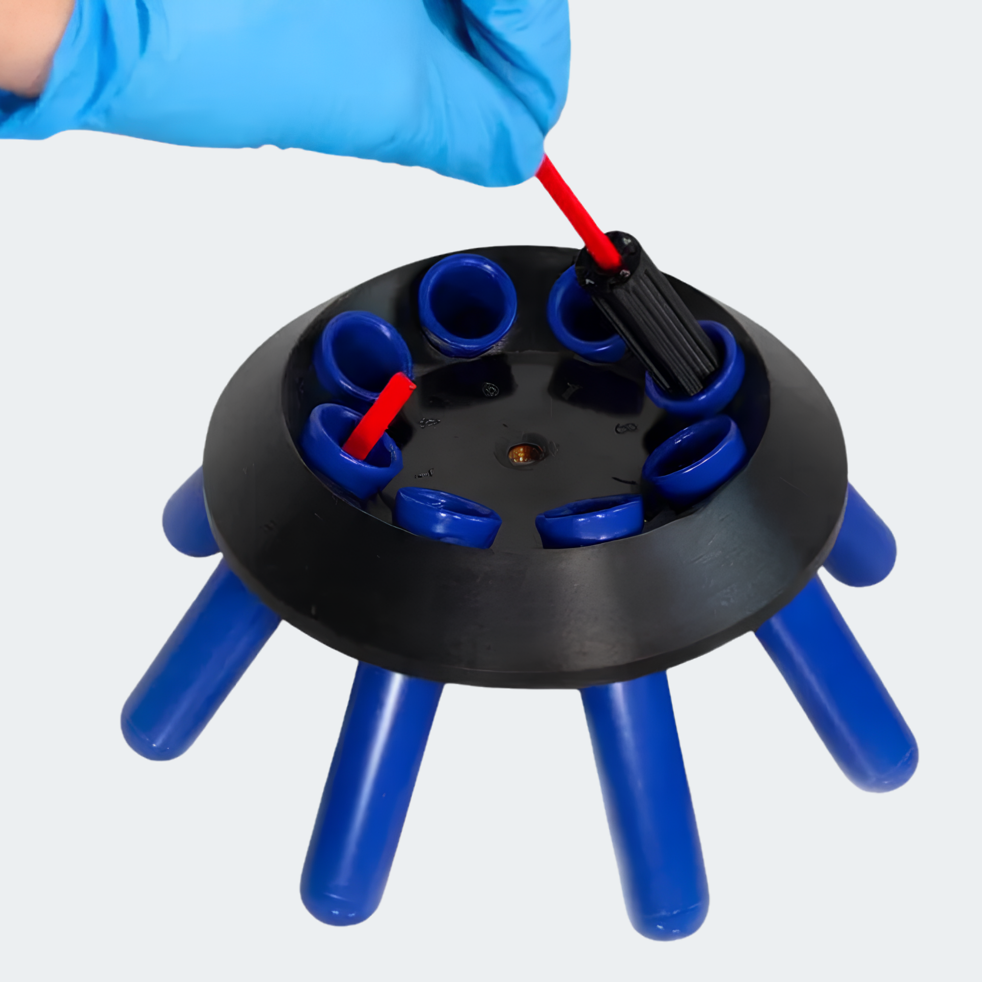 A blue-gloved hand inserts a red and black plastic accessory into one of the blue sockets on a black circular stand with blue legs, shown against a light gray background.