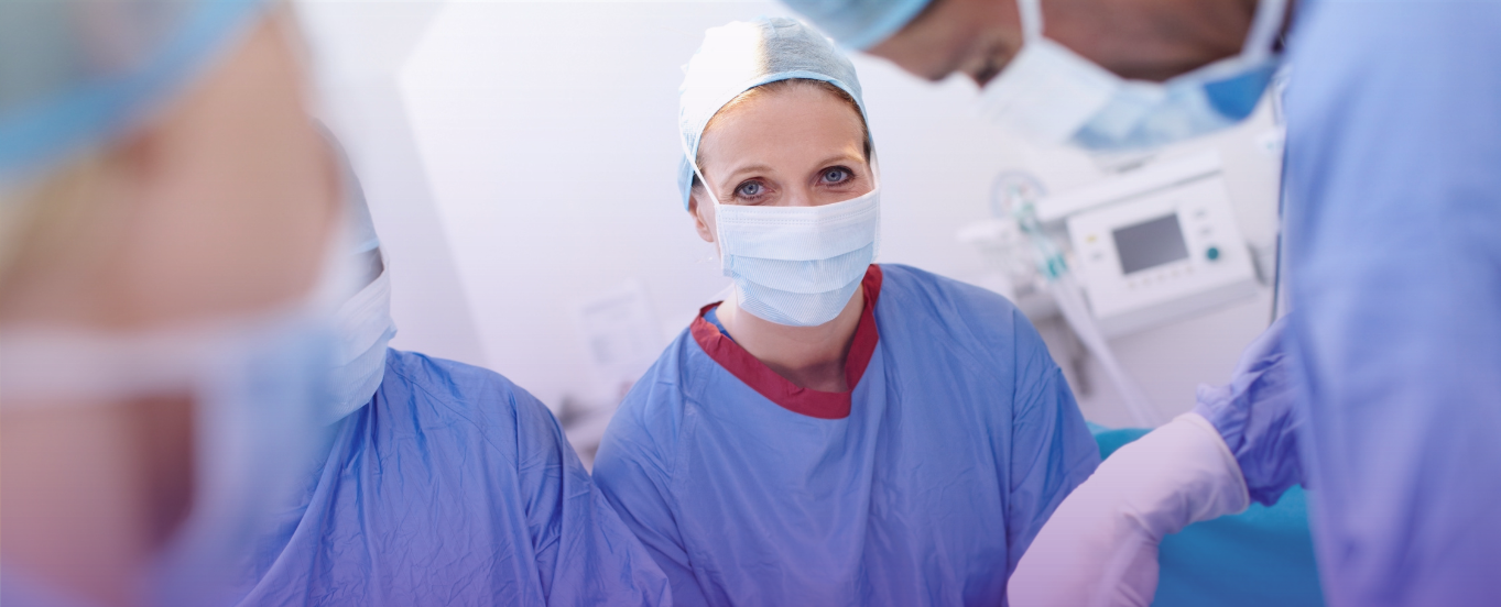 Three surgeons in an operating room, masked and gloved, with the female surgeon in the foreground looking forward, symbolizing a prepared and resilient medical team at the point of care.