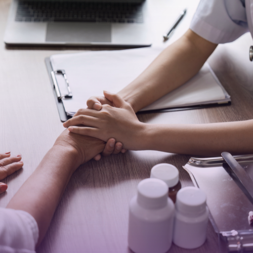 Close-up, overhead view of a medical professional gently holding the hand of an elderly patient across a desk, with a laptop, clipboard, and medicine bottles visible, symbolizing compassionate patient care and support.