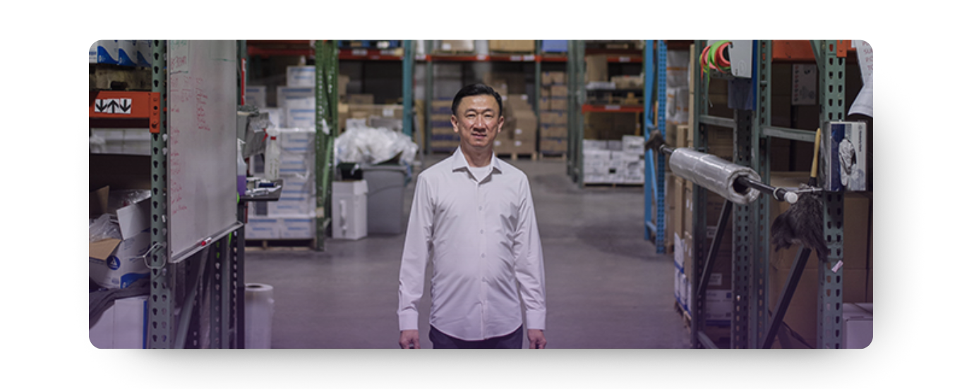Under HISTORY: A smiling Eric Liew, CEO of AOSS Medical Supply, stands confidently in a well-stocked warehouse aisle, looking directly at the camera. His presence amidst the inventory reflects his hands-on leadership and the company's foundational roots in efficient distribution.