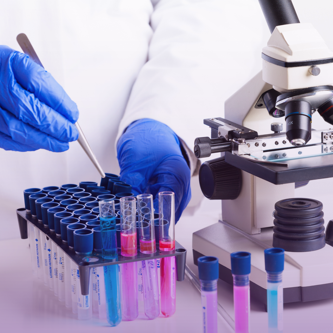 Gloved hands in a lab coat using forceps to adjust colorful liquid samples in test tubes on a rack, with a microscope in the background, indicating laboratory work.