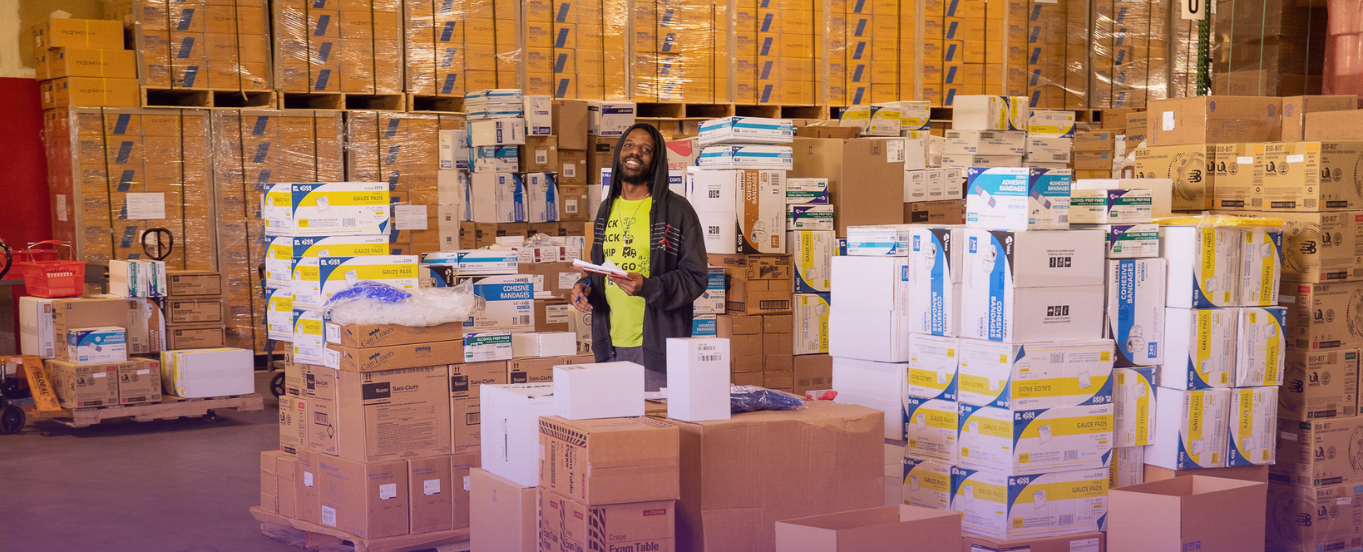A male warehouse worker with dreadlocks, wearing a neon yellow shirt and dark jacket, holding a tablet or scanner, stands amidst tall stacks of medical supply boxes in a large, busy warehouse.